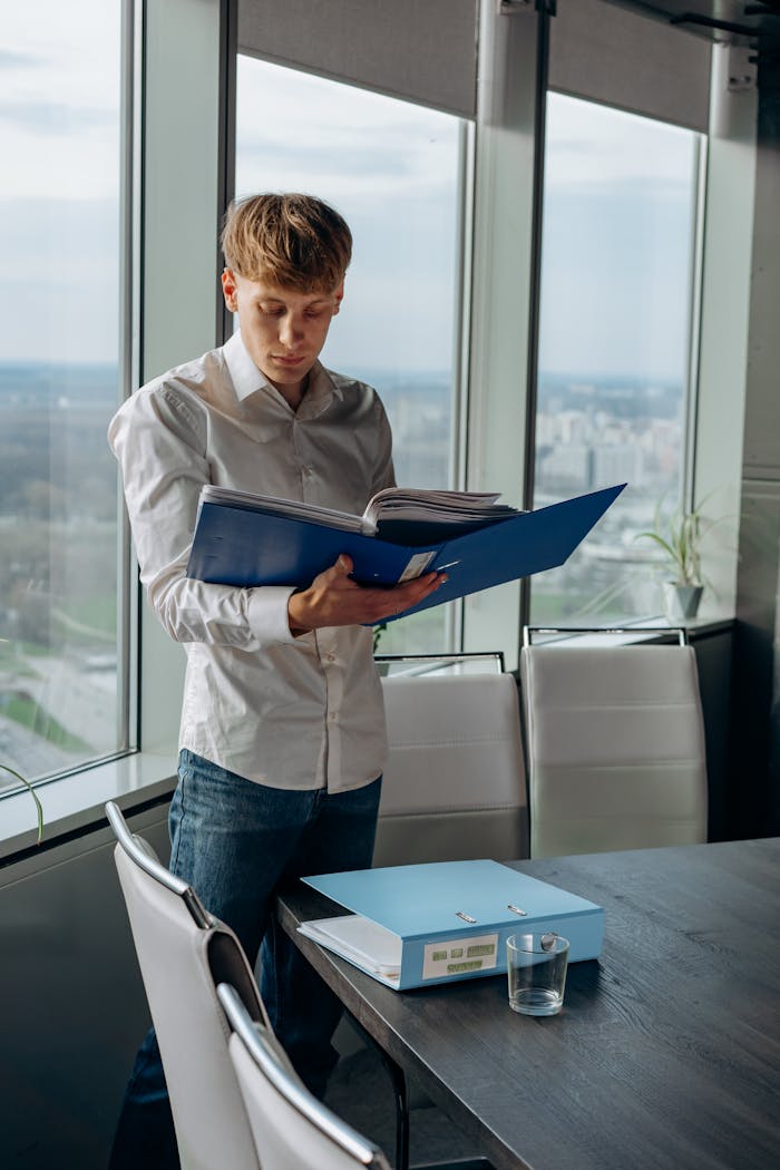 Young professional reading documents by a window with an urban view.